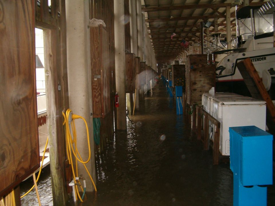 Boats docked in a flooded marina with wooden pilings and yellow hoses
