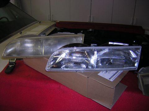 1992 Ford Crown Victoria - Two composite headlight assemblies with clear lenses and chrome reflectors, one resting on a cardboard shipping box and the other placed on…