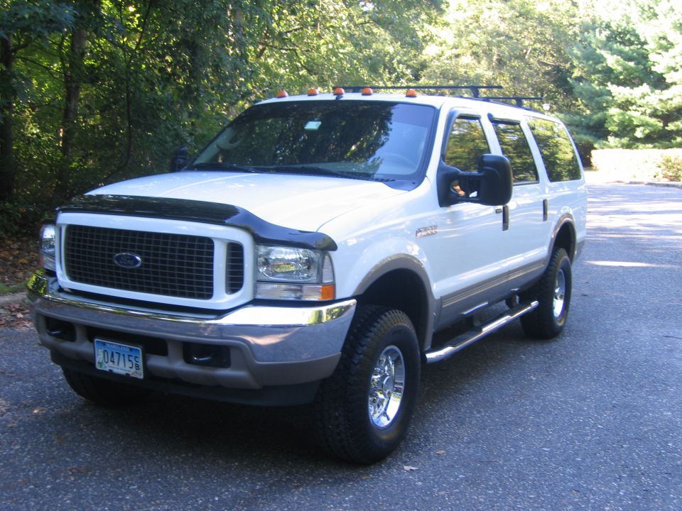 White Ford Excursion SUV with tan lower trim, chrome wheels and hood deflector, parked on a tree-lined driveway