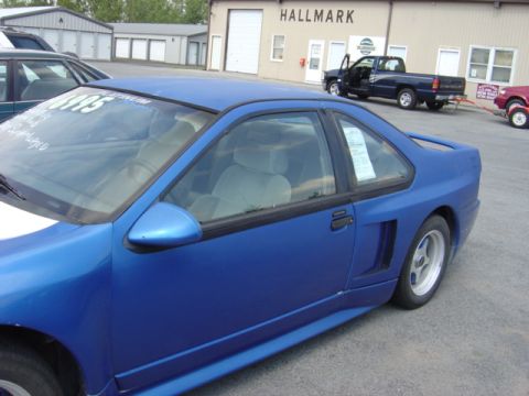 1998 Ford Taurus - A blue coupe is parked next to a building with a Hallmark sign, with other vehicles in the background.