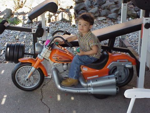 1989 Ford Bronco - A young boy sits on an orange toy motorcycle next to a weight rack outdoors.