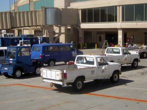 1995 Ford Bronco - Multiple vehicles are parked on an airport tarmac near a jet bridge and terminal building.