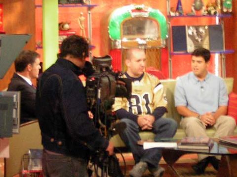 1995 Ford Bronco - A television studio set is prepared for filming, a camera operator, two guests seated on a couch, and a vintage jukebox in the background.