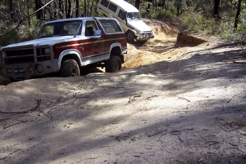 1982 Ford Bronco - Two off-road vehicles tackle a challenging sandy track in a wooded area.