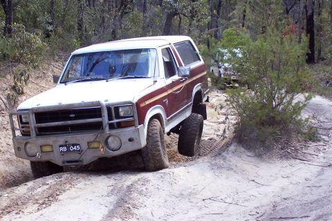 1982 Ford Bronco - A white and maroon Ford Bronco navigates a sandy, wooded trail with another vehicle visible in the background.