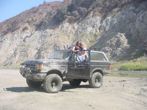 1988 Ford Bronco - This heavily muddied 1987 Ford Bronco, with its door ajar and two occupants visible, sits in a dusty, off-road environment.