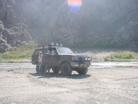1988 Ford Bronco - 1988 Ford Bronco parked in a dirt lot with a rocky hillside behind it. Two people are near the vehicle.