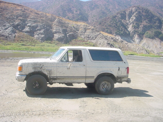 Mud-caked white full-size Ford Bronco parked side-on in a dry riverbed below brown hills