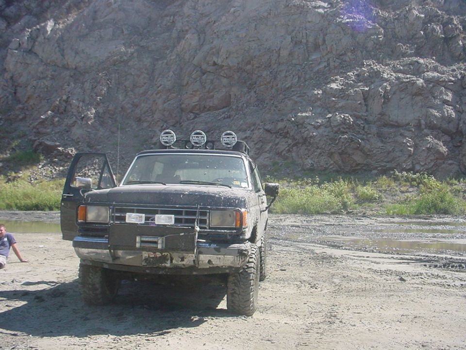 Dark-colored Ford Bronco II with off-road lights on roof, parked on dirt with a person sitting nearby