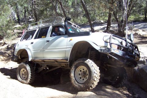 1982 Ford Bronco - A white SUV is shown off-roading on a dirt path with trees in the background.
