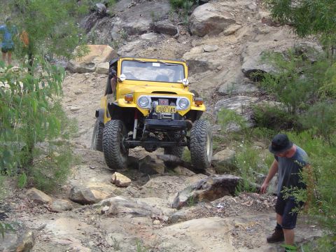 1982 Ford Bronco - A yellow Toyota Land Cruiser navigates a challenging rocky trail with a person watching nearby.