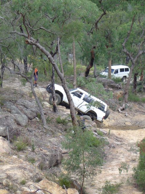 1982 Ford Bronco - Two white SUVs tackle a challenging off-road course amidst trees and rocks.