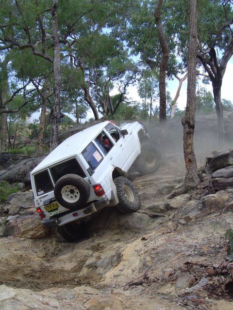 1982 Ford Bronco - An off-road vehicle navigates a challenging rocky incline, kicking up dust as it climbs.