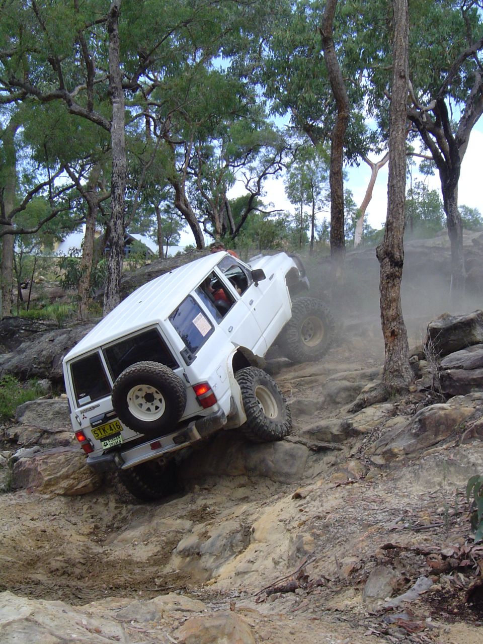 White SUV is going up a steep, rocky hill with trees around. It has a spare tire on the back