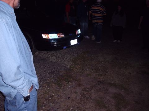 1998 Ford Taurus - A group of people gather around a black sedan at night, illuminated by its headlights.