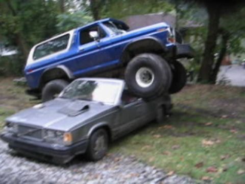 1979 Ford Bronco - A blue Ford Bronco is parked on top of a grey sedan, creating a precarious scene.