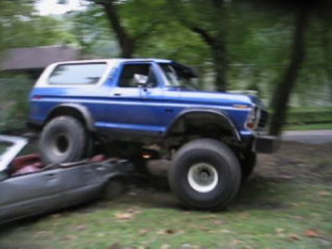 1979 Ford Bronco - A lifted blue and white SUV is shown driving over a dark car in an outdoor setting.
