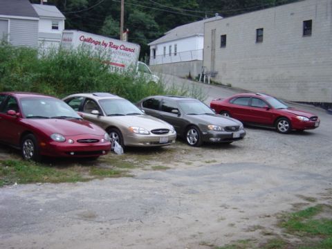 1998 Ford Taurus - Several sedans are parked in a gravel lot next to a building and some bushes.