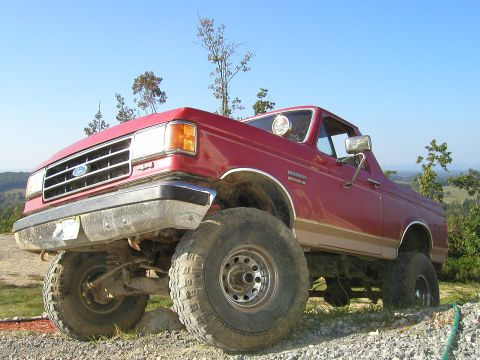 1989 Ford Bronco - This lifted red Ford Bronco is parked on a gravelly surface with trees and hills in the background.