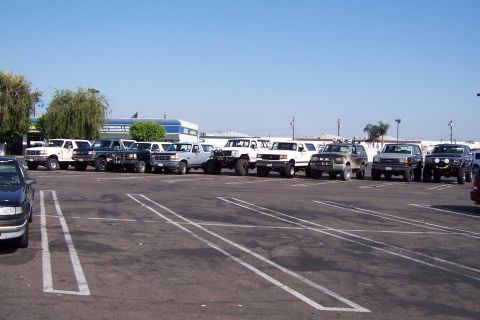 1990 Ford Bronco - Multiple Ford Broncos and trucks are parked in a lot under a clear blue sky.