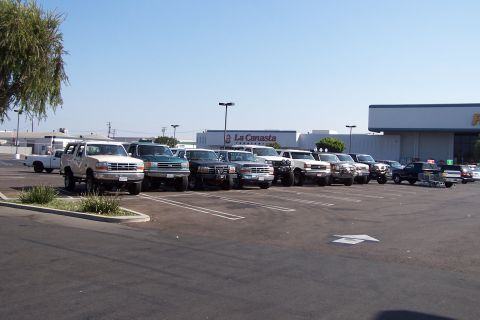 1990 Ford Bronco - A line of trucks and SUVs are parked in a lot on a clear day, with a store in the background.