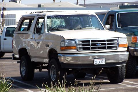 1990 Ford Bronco - A tan 1990 Ford Bronco with a cracked windshield and visible damage to its side is parked next to another vehicle.