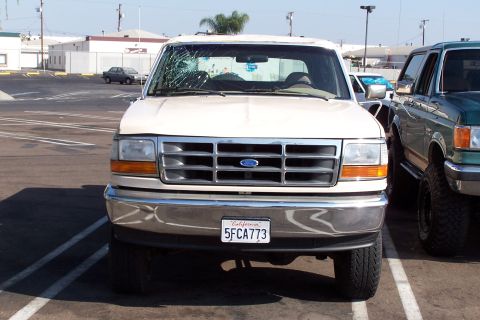 1990 Ford Bronco - A white Ford Bronco with a cracked windshield is parked next to a green SUV in a parking lot.