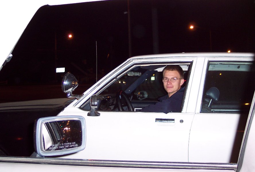 White sedan at night with smiling driver visible through open window, chrome side mirror in foreground