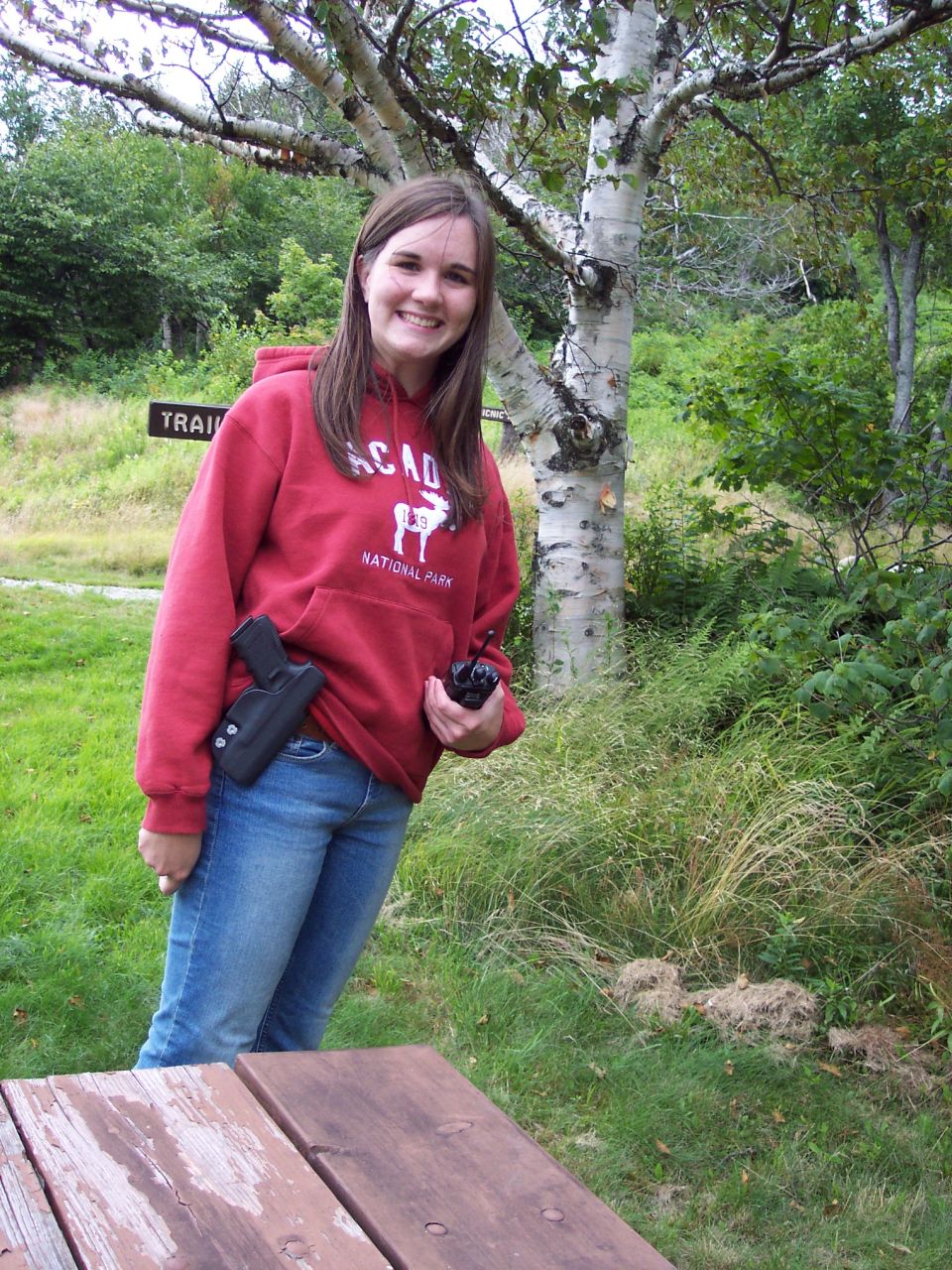 Woman in red Acadia National Park hoodie holding a radio, standing by a birch tree and picnic table