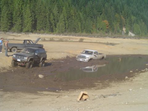 1993 Ford Bronco - Several off-road vehicles are stuck in a muddy area with trees in the background.