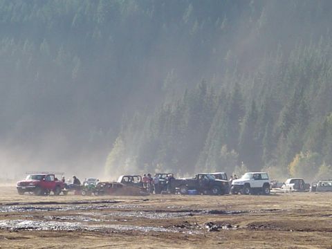 1993 Ford Bronco - A collection of off-road vehicles are parked in a muddy field, with a dense forest visible behind them.