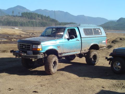 1993 Ford Bronco - Modified blue pickup truck with a camper shell on a dirt road, with mountains in the background.
