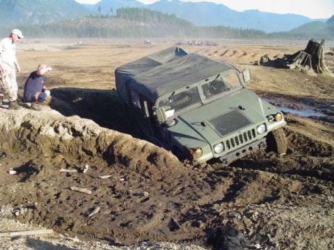 1993 Ford Bronco - A military vehicle is stuck in a muddy ditch, with two individuals looking on from the edge.