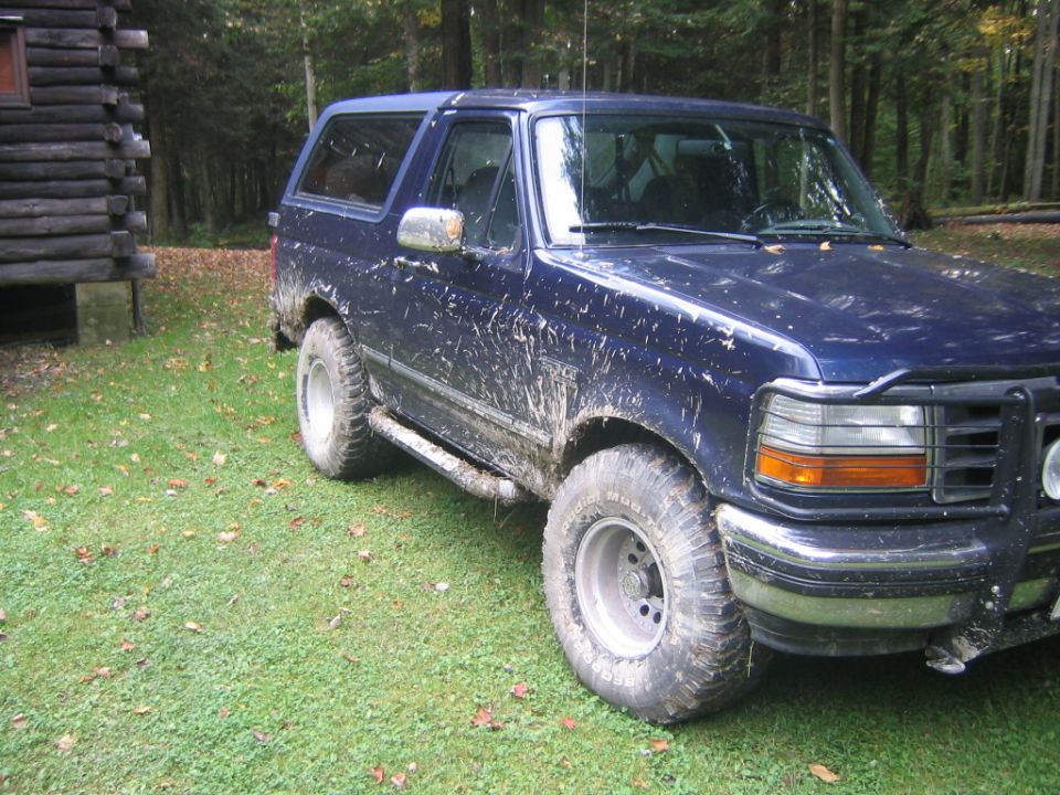 Blue Ford Bronco covered in mud, parked on grass next to a log cabin