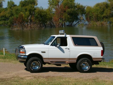1996 Ford Bronco - A white Ford Bronco with a tan top is parked on a dirt path next to a body of water.