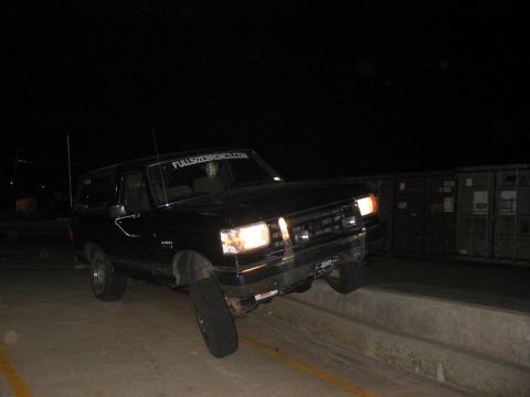 1988 Ford Bronco - A black SUV is positioned precariously on a concrete barrier at night, with a website sticker visible on its windshield.