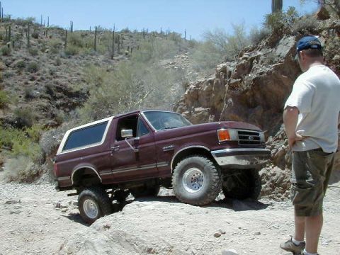 1990 Ford Bronco - A maroon 1990 Ford Bronco navigates a challenging rocky terrain under a clear blue sky.