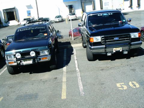 1989 Ford Bronco - Two trucks parked in a lot. One is a black Ford Bronco, the other is a dark SUV with a "Pathfinder" sticker.