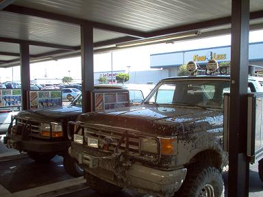 1988 Ford Bronco - Two Ford Broncos are parked under a covered area, one heavily covered in mud.
