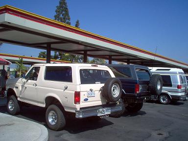 1988 Ford Bronco - Three vehicles, including a tan Ford Bronco, are parked under a covered structure on a sunny day.