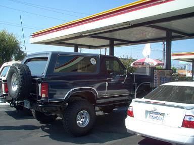 1988 Ford Bronco - A dark blue 1988 Ford Bronco is parked at a gas station, showing its rear spare tire and lifted suspension.