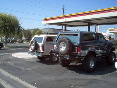1988 Ford Bronco - Two Ford Bronco SUVs are parked at a gas station, viewed from the rear.