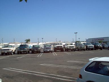 1988 Ford Bronco - A variety of trucks and SUVs are parked in a lot, with a building in the background.