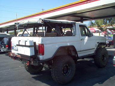 1988 Ford Bronco - Modified white Ford Bronco with off-road tires and roll cage, seen from the rear three-quarter view.