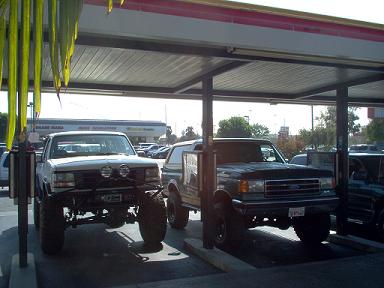 1988 Ford Bronco - Two Ford Broncos are parked side-by-side under a covered structure, possibly a carport or gas station awning.