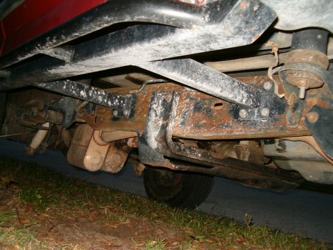 1991 Ford Bronco - The rusty undercarriage of a red vehicle is visible from a low angle, showing the frame and exhaust components.