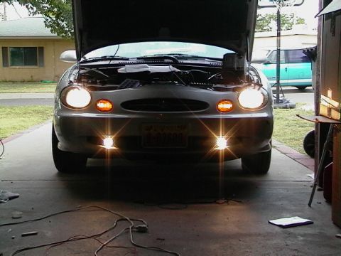 1999 Ford Taurus - Front view of a 1999 Ford Taurus with the hood up, headlights and fog lights illuminated.