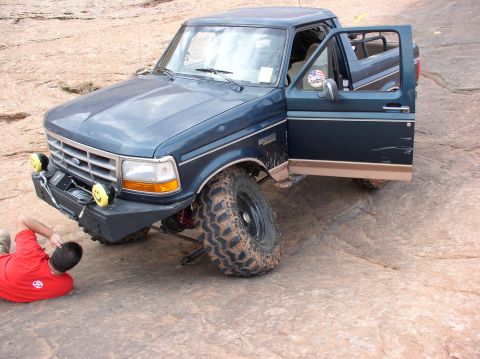 1993 Ford Bronco - A person works on the front tire of a blue 1993 Ford Bronco parked on rocky terrain.