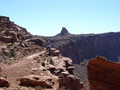 1993 Ford Bronco - An off-road vehicle navigates a dirt road in a vast canyon, with dramatic rock formations and a distant peak.