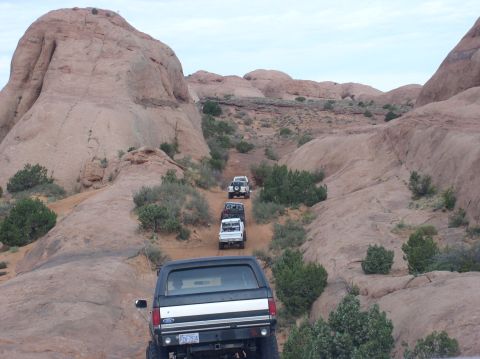 1993 Ford Bronco - A convoy of off-road vehicles navigates a challenging desert trail, with the lead Bronco climbing a steep incline.